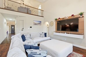 A spacious living room with a large flat-screen TV, a white sofa, a white armchair, and a wooden staircase. - 5BD, 4BA Entertainment Hub - Playground, Cinema, Gym (Frisco)