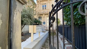 A gated entrance with a metal gate, a pathway leading to a building with a balcony, and a small garden area with white pebbles and plants. - Charming Stay in the Heart of Vittel (Vittel)
