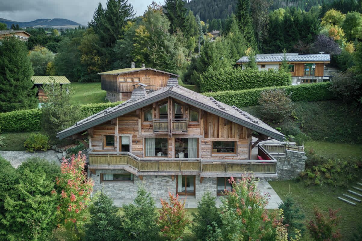 A wooden chalet with a stone foundation, multiple balconies, and a view of a lake surrounded by greenery.