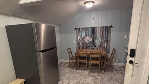 Kompakti keittiö, jossa on sisäänrakennettu ruostumattomasta teräksestä valmistettu jääkaappi, ruokapöytä tuoleineen ja puinen jakkara.