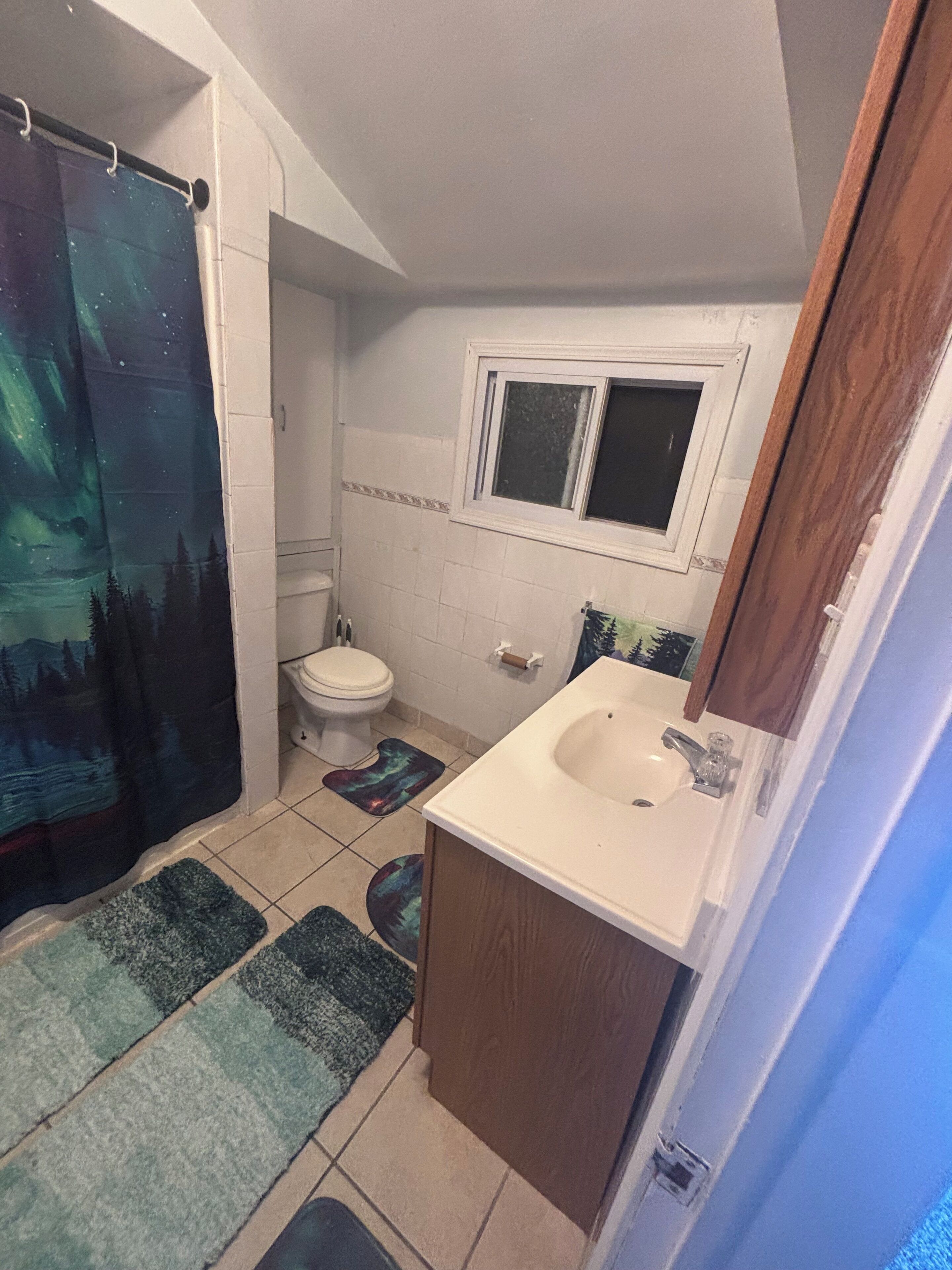 A small bathroom with a toilet, sink, and window. There are two rugs on the floor.