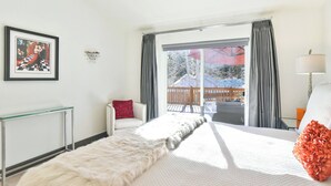 Une chambre d’hôtel avec un grand lit, une table en verre, une chaise rouge, un plaid en fausse fourrure blanche, un coussin en forme de fleur rouge, et une vue sur une terrasse en bois et des arbres à l’extérieur.