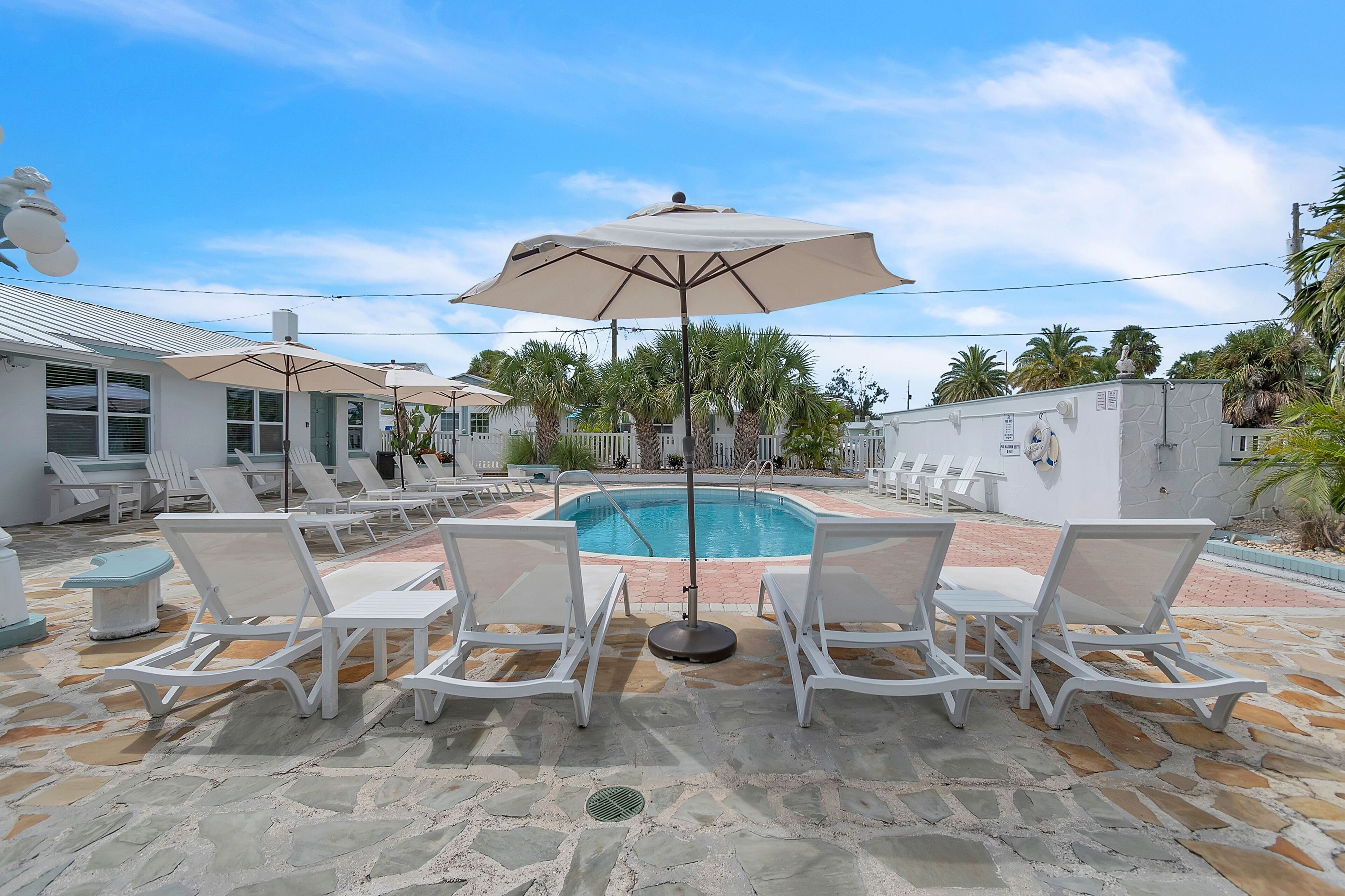 A poolside area with white lounge chairs and umbrellas, a stone patio, and a clear blue sky.