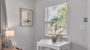 Una habitación con una mesa blanca, una obra de arte enmarcada, una ventana con persianas y una lámpara en una mesita de noche.