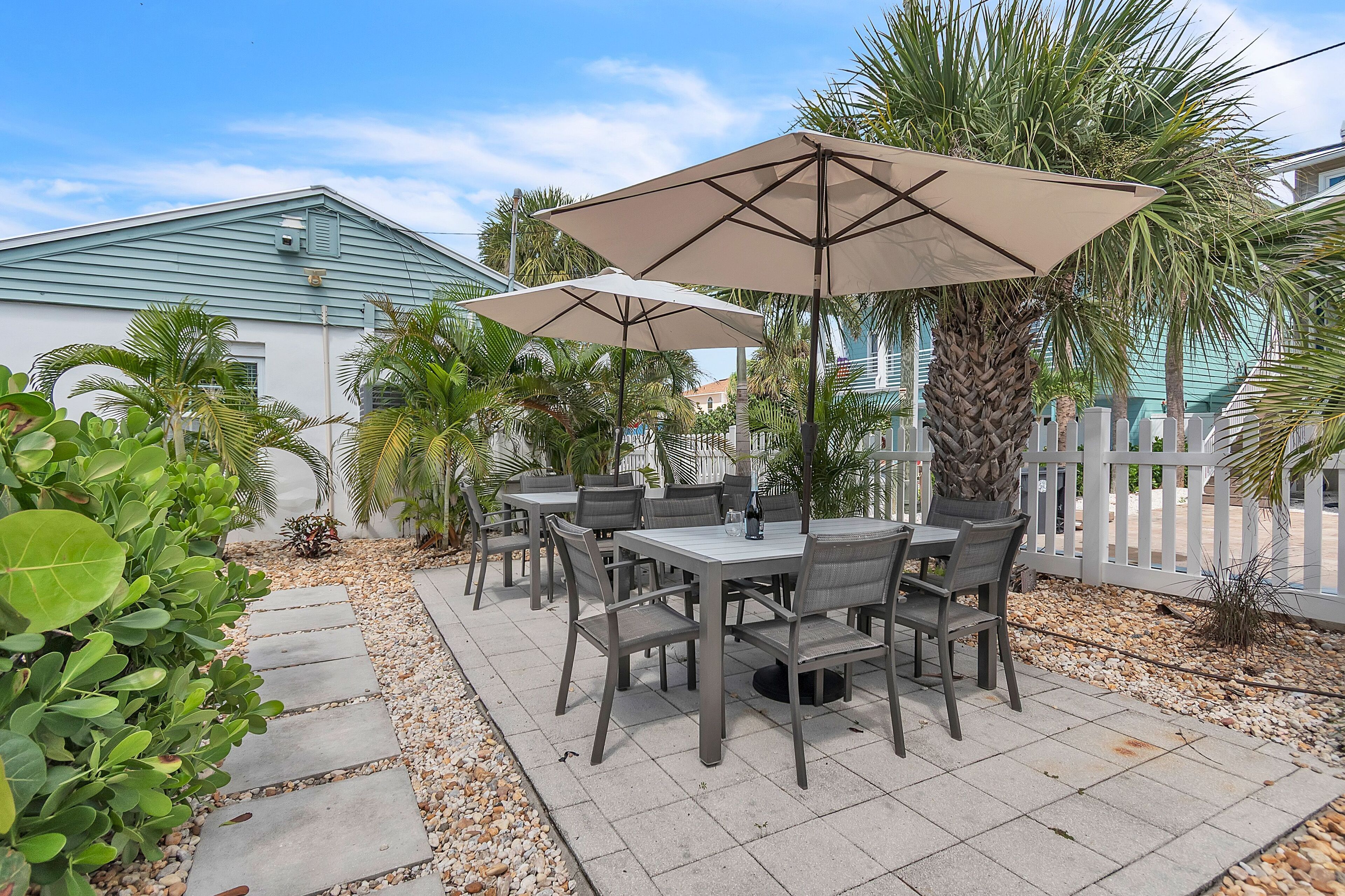 An outdoor patio with a table and chairs, surrounded by palm trees and a white picket fence.