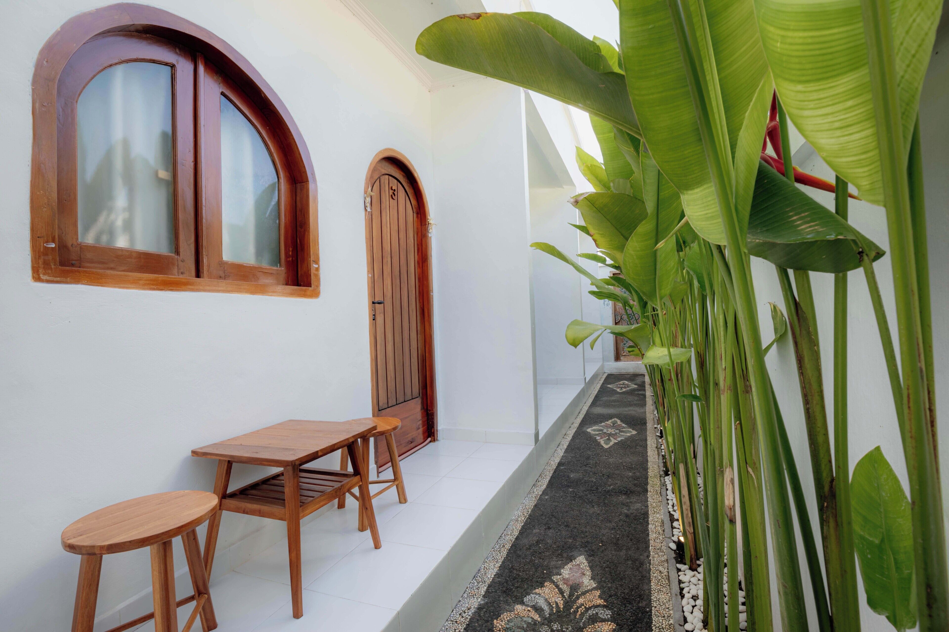Un pasillo estrecho con una mesa y sillas de madera, una puerta de madera y grandes plantas verdes a lo largo de la pared.