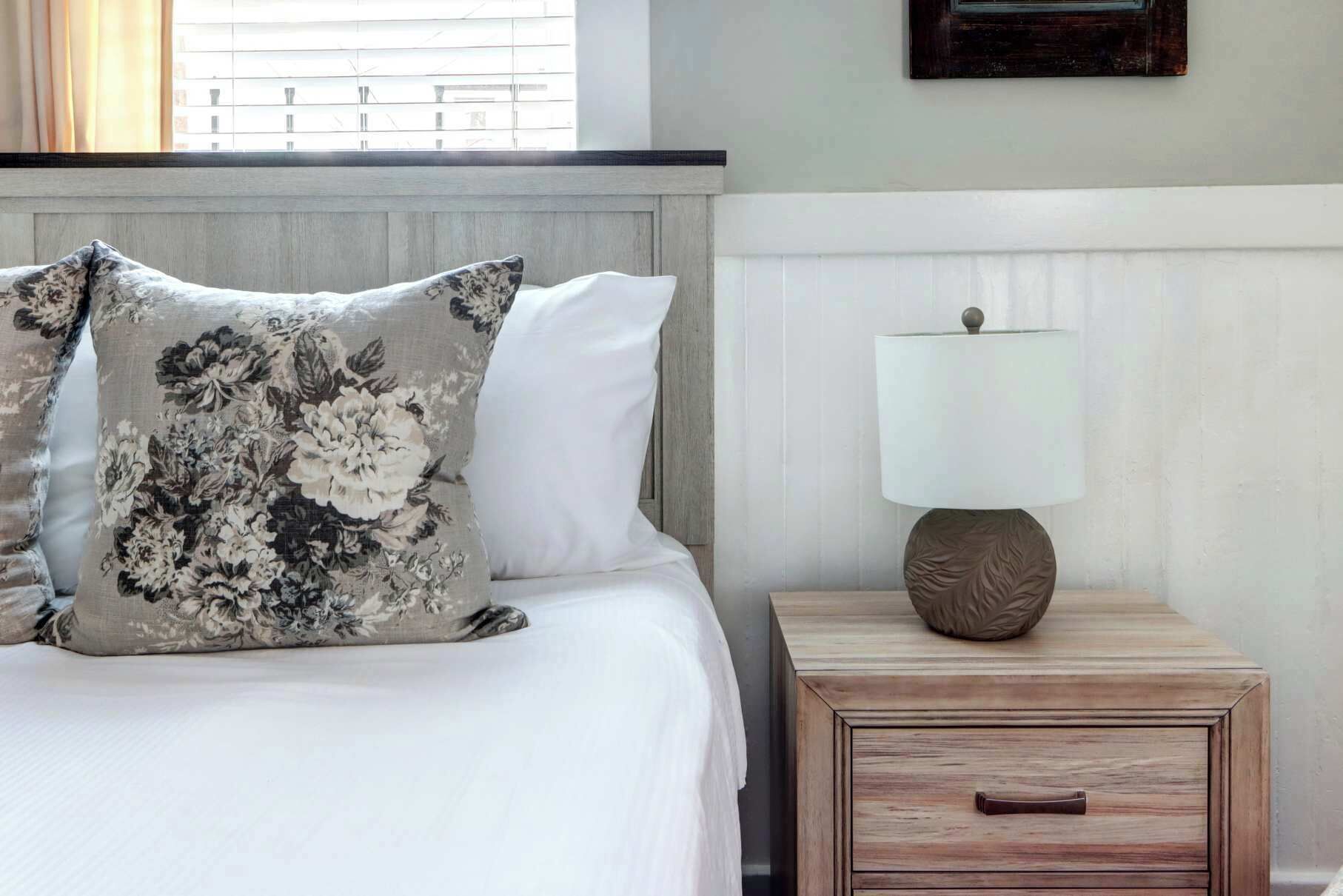 A neatly made bed with floral pillows, a bedside lamp, and a wooden nightstand with a drawer.