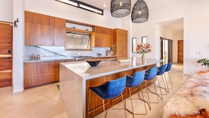 A modern kitchen with wooden cabinets, a large island with bar stools, pendant lighting, and a view to the living area. - Villa Damiana by The Agency Baja (El Pescadero)