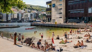 A waterfront area with a dock, people swimming and relaxing on the sand, and buildings in the background. - Brand New & High-End, 3 Bedrooms 2 Bathrooms, Central Apartment (Bergen)