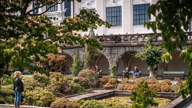 A garden with benches, a fountain, and a building with arched windows in the background. - Brand New & High-End, 3 Bedrooms 2 Bathrooms, Central Apartment (Bergen)