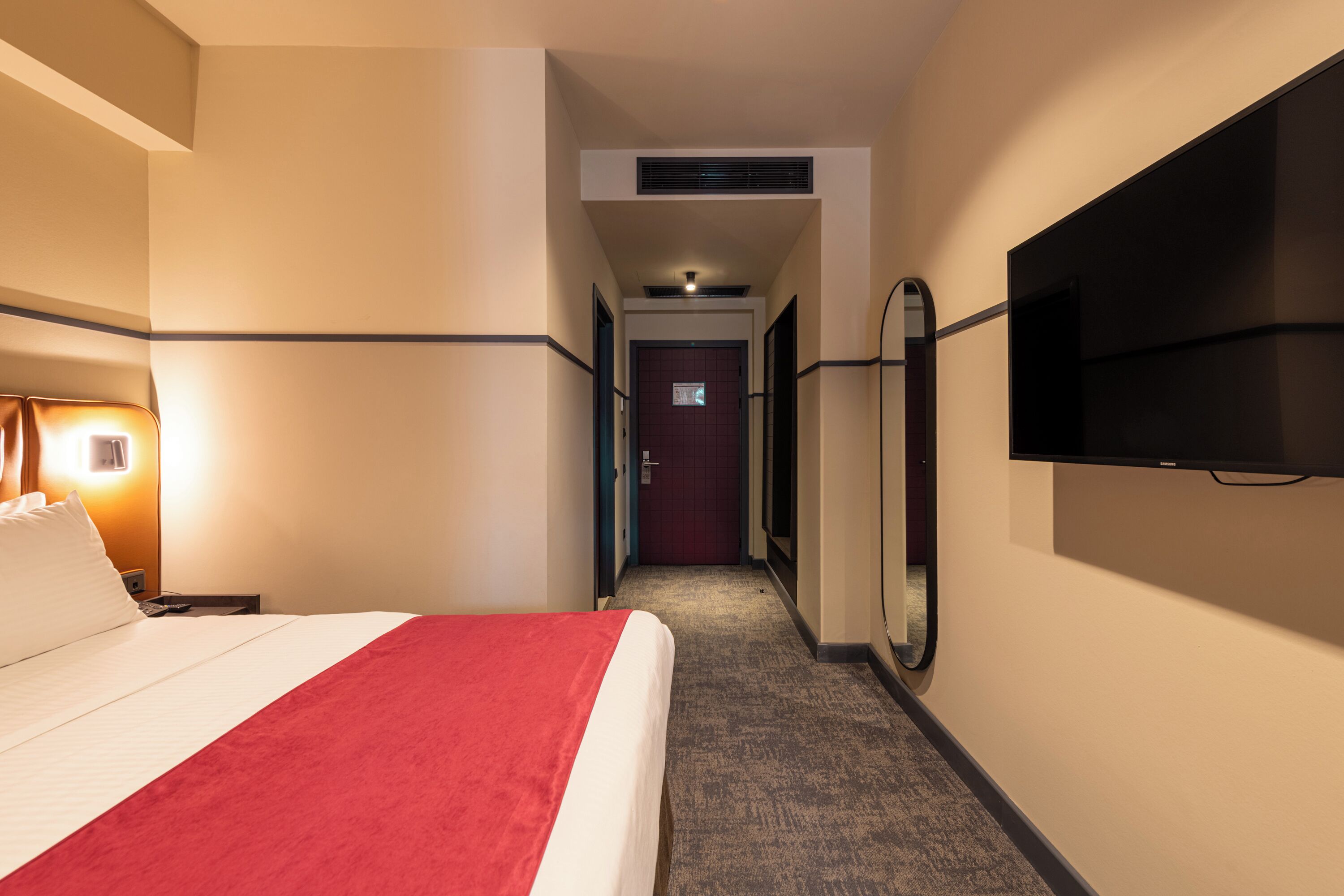 Habitació d'hotel amb un llit, televisió, mirall i un passadís que condueix a una porta.