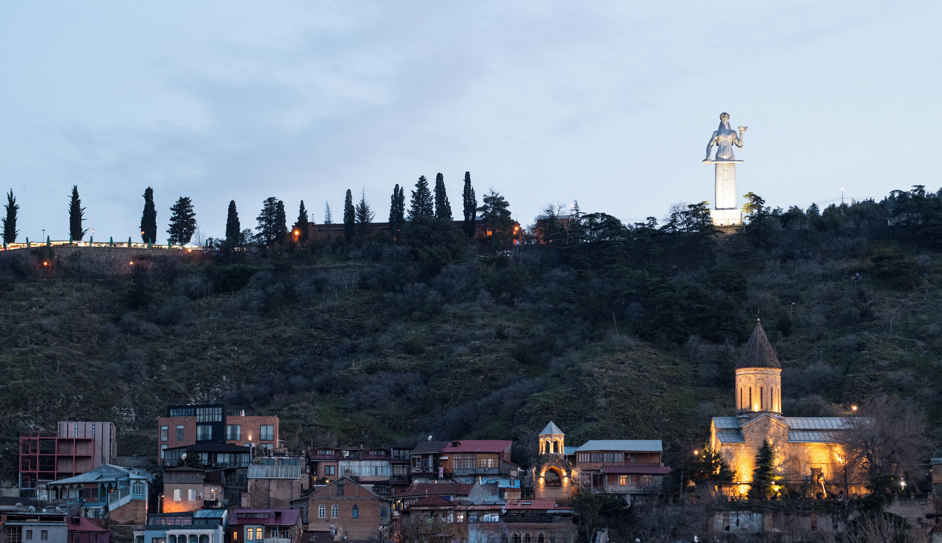 Un pueblo con una estatua destacada en una colina, una iglesia con un campanario y varios edificios en la base.