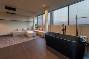 Ein modernes Badezimmer mit freistehender Badewanne, einer Sitzecke mit Sofa und Hocker sowie großen Fenstern mit Jalousien.