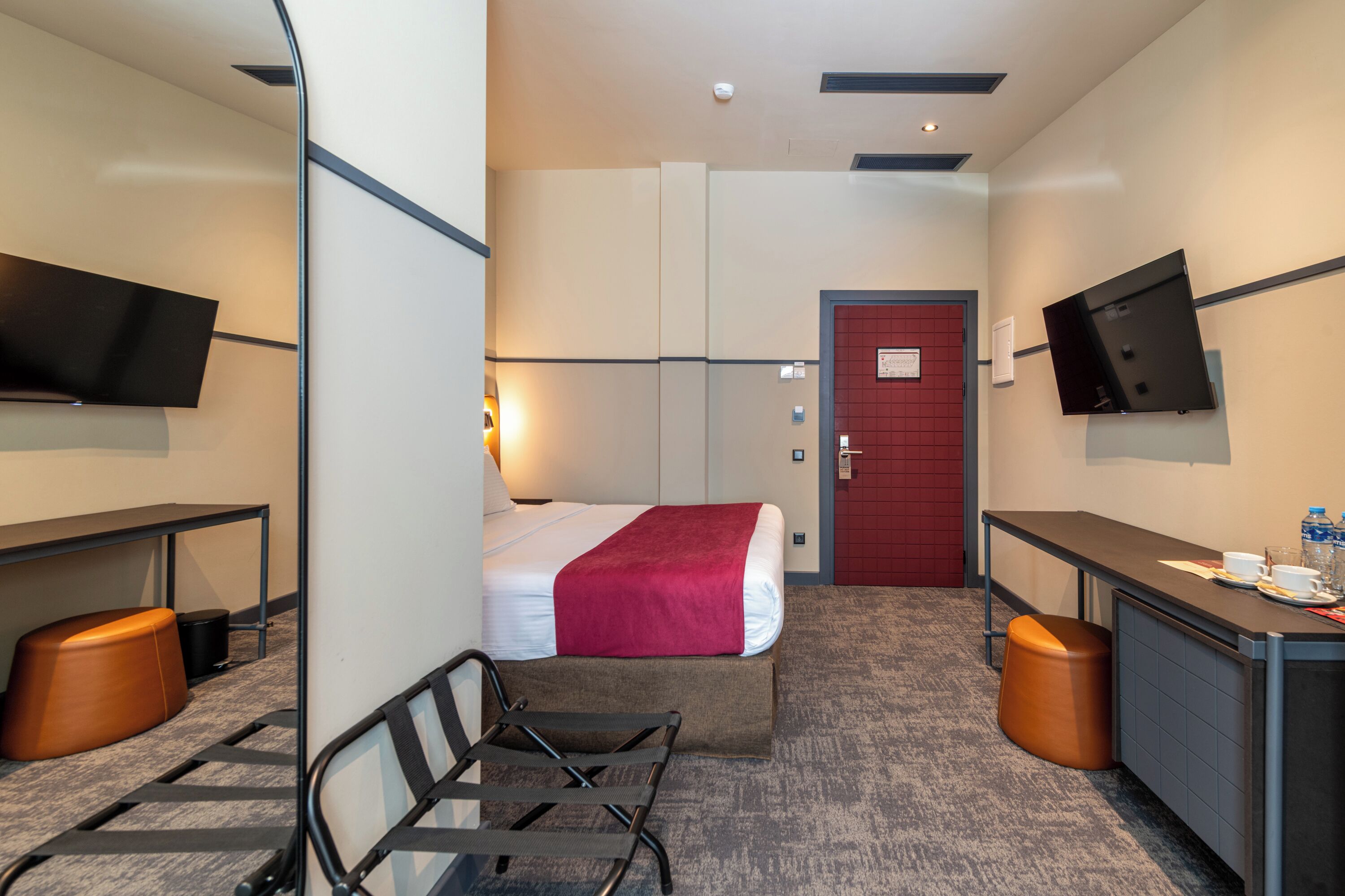 A hotel room with a bed, a desk, a chair, a television, and a red door.