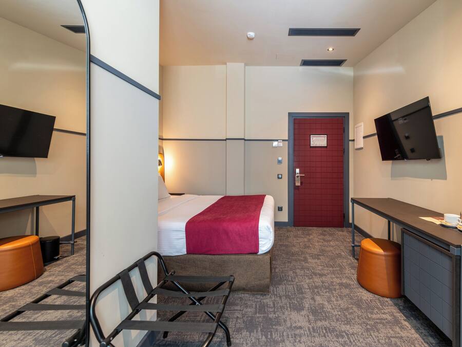A hotel room with a bed, a desk, a chair, a television, and a red door.