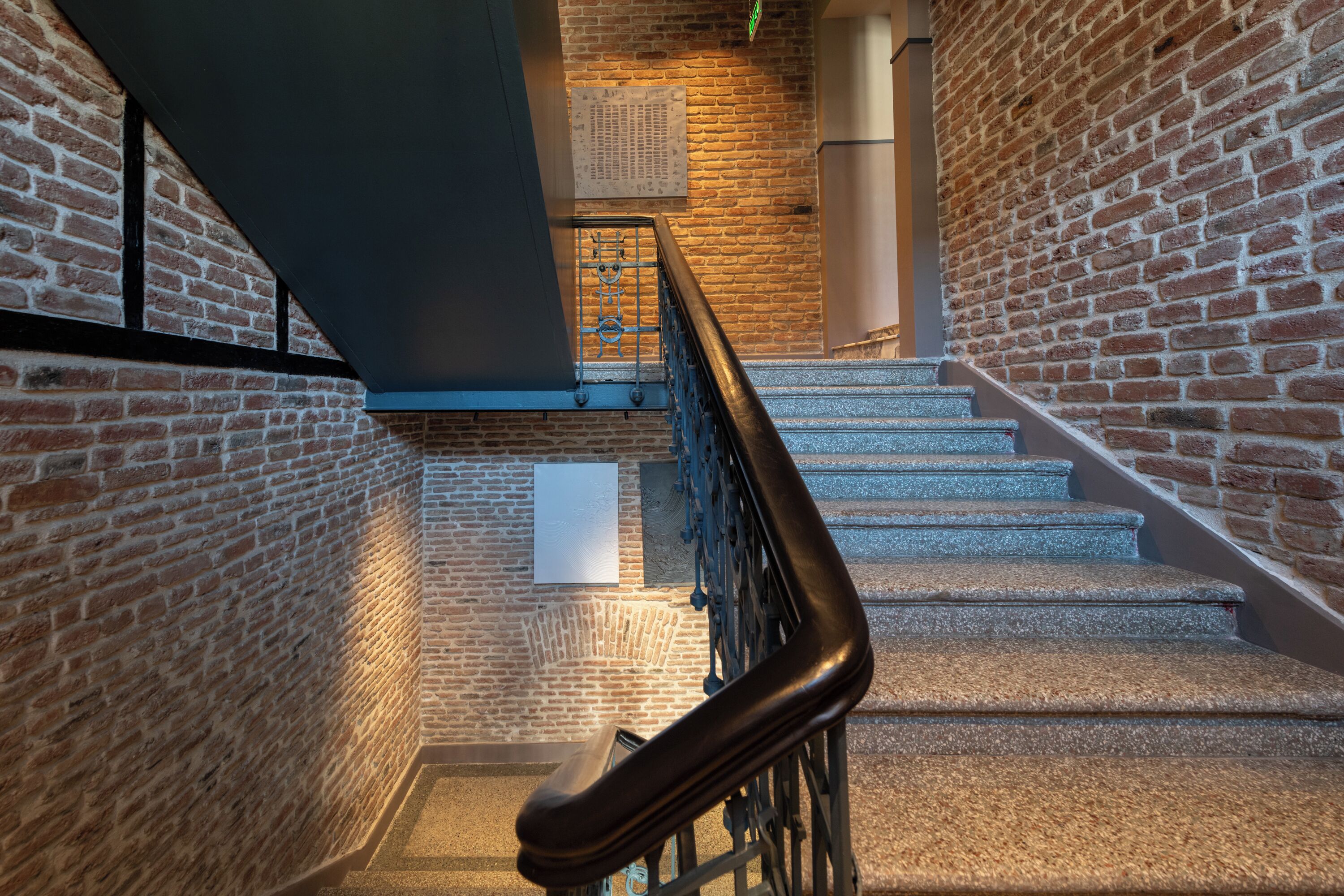 A staircase with a metal handrail, brick walls, and a ceiling vent.