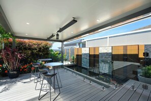 Une terrasse extérieure dotée d’une plateforme en bois, de chaises en métal et d’une balustrade en verre, offrant une vue sur un jardin et un bâtiment.