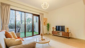 Ein geräumiges Wohnzimmer mit einer Couch, einem Couchtisch, einem Fernseher auf einem Holzboden und einer Schiebetür zu einem Balkon mit Blick auf Gebäude und Grünflächen.