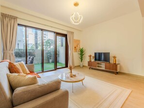 En rummelig stue med sofa, sofabord, fjernsyn på et træstel og en skydedør, der fører ud til en balkon med udsigt til bygninger og grønne områder.