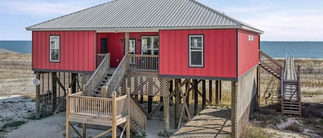 A red wooden house with a metal roof, a wooden staircase, and a sandy beach in the background.