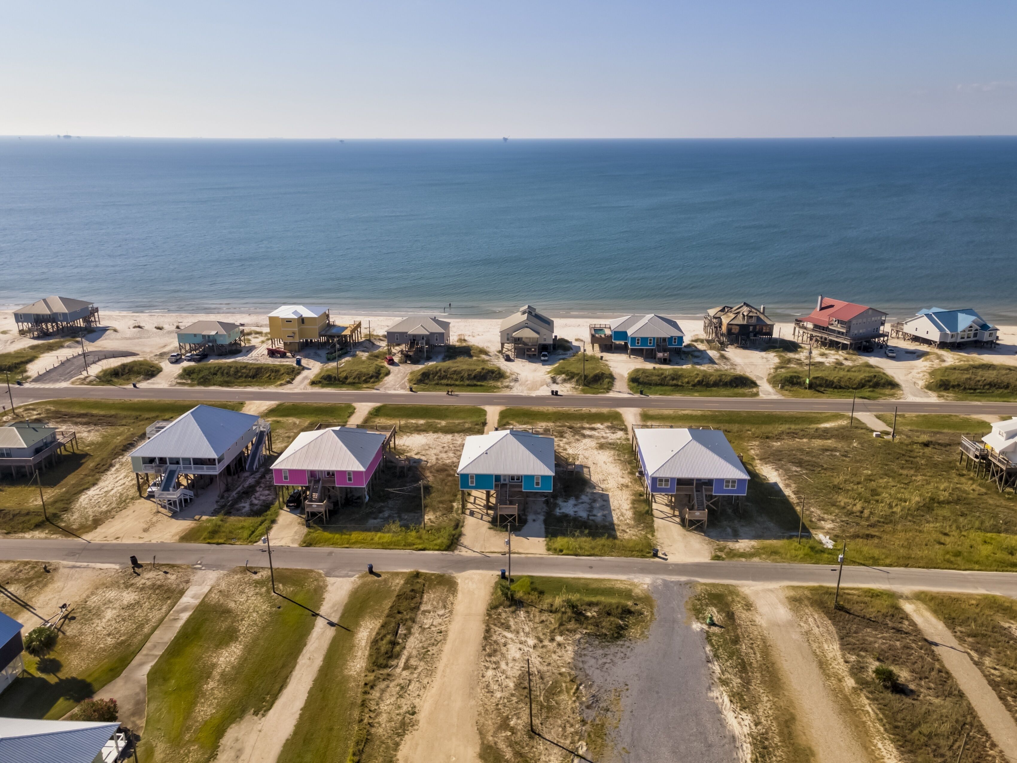 An aerial view of a coastal residential area with houses on stilts, a beach, and a road.