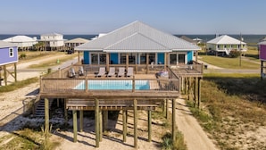 A beachfront house with a pool on a wooden deck, surrounded by sandy terrain and other houses.