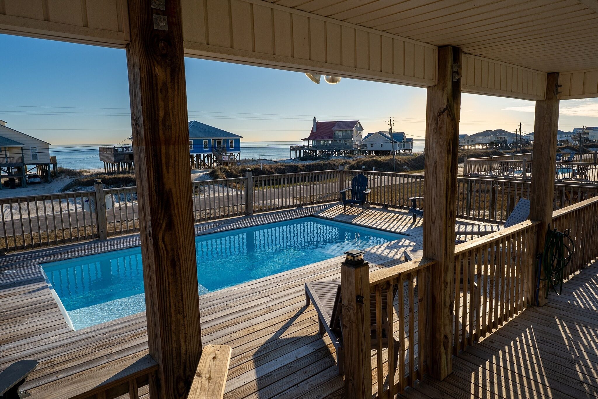 Eine Holzterrasse mit Pool, mit Blick auf einen Strand mit Häusern und einem klaren Himmel.