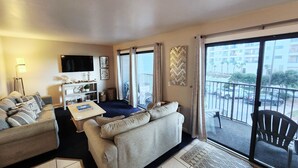 Een woonkamer met een bank, een salontafel en een televisie. Er is een balkon met uitzicht op de straat en de gebouwen.