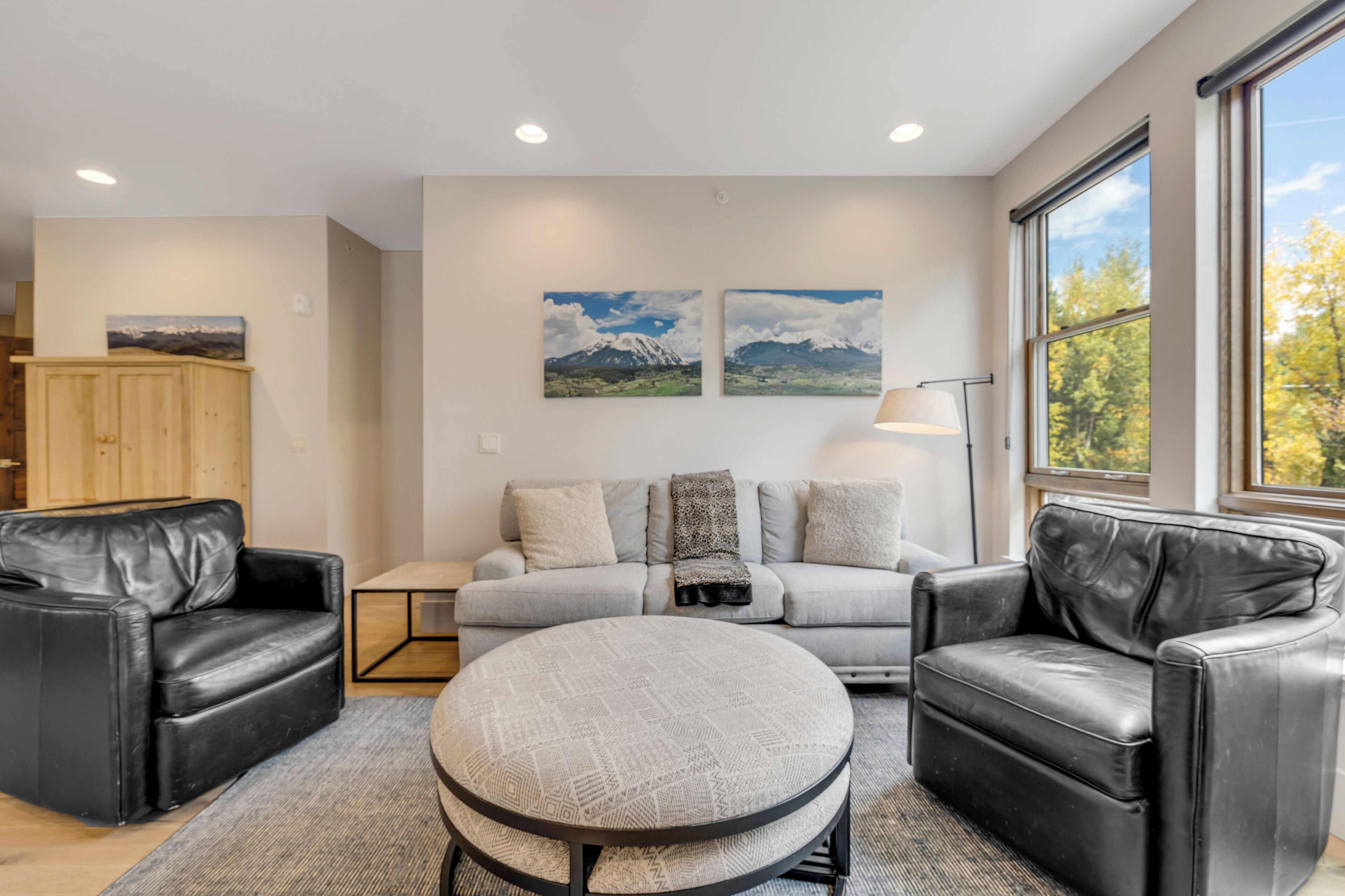 A modern living room with a grey sofa, two black leather armchairs, a round coffee table, and a wooden cabinet. There are two framed landscape paintings on the wall and a floor lamp.