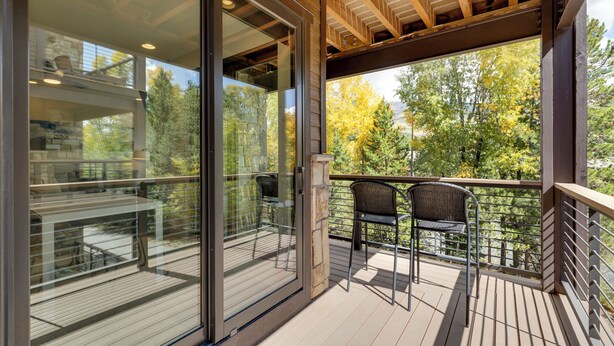 Um deck de madeira com portas de vidro que dão para uma varanda, móveis de exterior e vista para árvores.