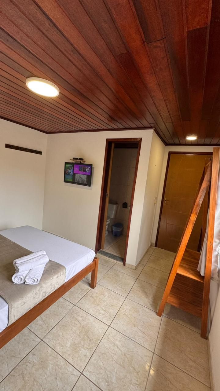 A bedroom with a wooden ceiling, a bed, a small TV on the wall, and a bathroom visible through an open door.