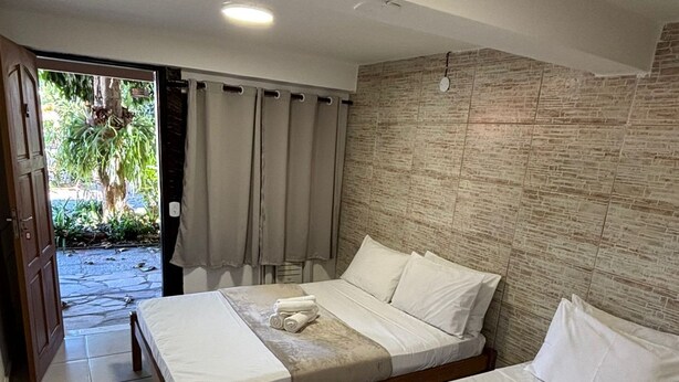 A hotel room with two beds, a wooden headboard, a stone-textured wall, and a view of a garden through an open door. - Go Hotelaria (Búzios)