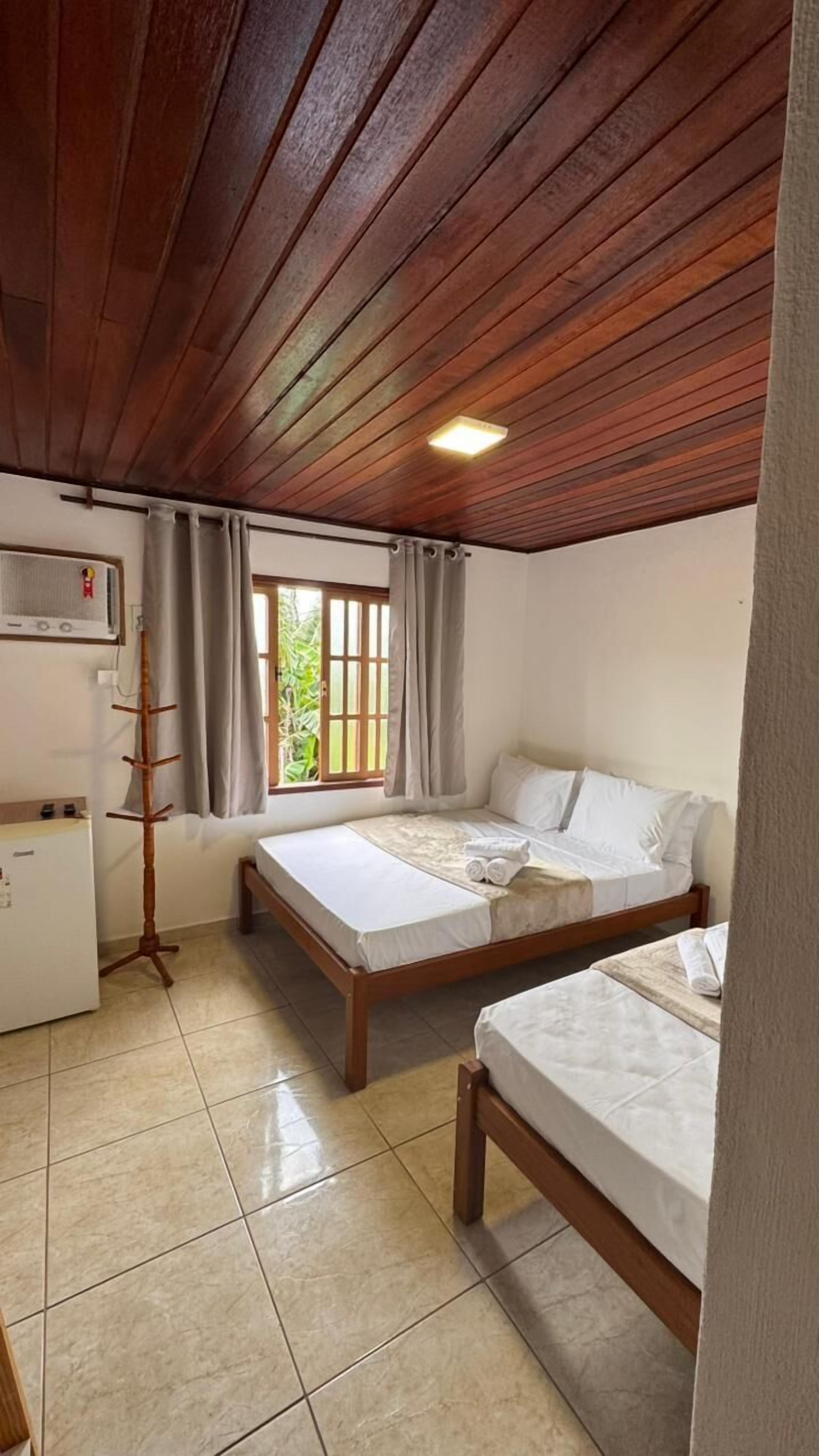 A room with two beds, a wooden ceiling, a window with curtains, a radiator, and a tiled floor.
