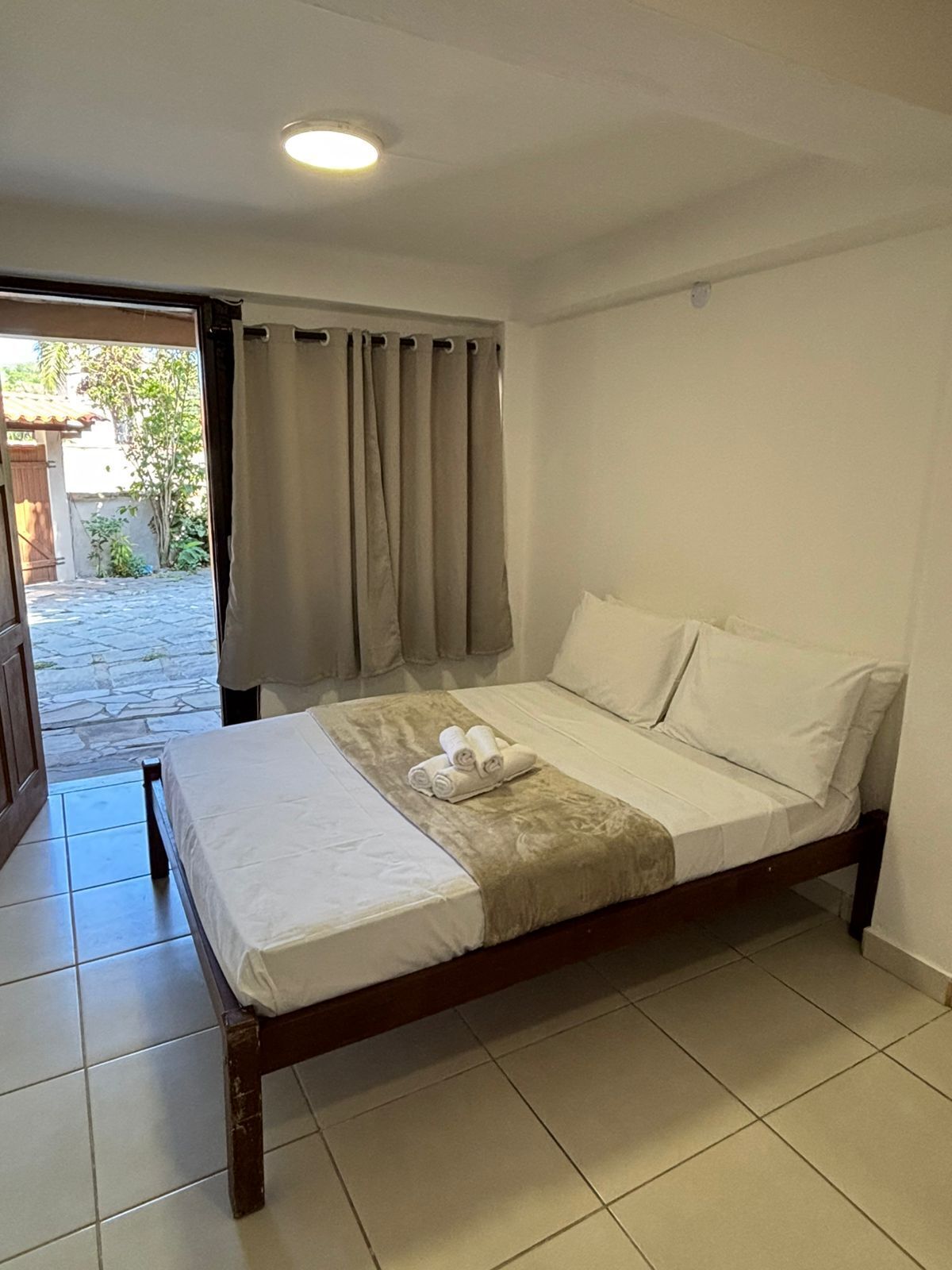 Un letto singolo con struttura in legno, biancheria da letto bianca e una coperta beige. Si vede una finestra con tende beige e una porta che dà su un'area esterna con un vialetto in pietra.