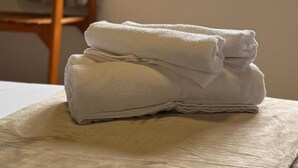 A neatly folded white towel on a bed with a wooden headboard, a window with vertical blinds, and a painting on the wall. - Go Hotelaria (Búzios)
