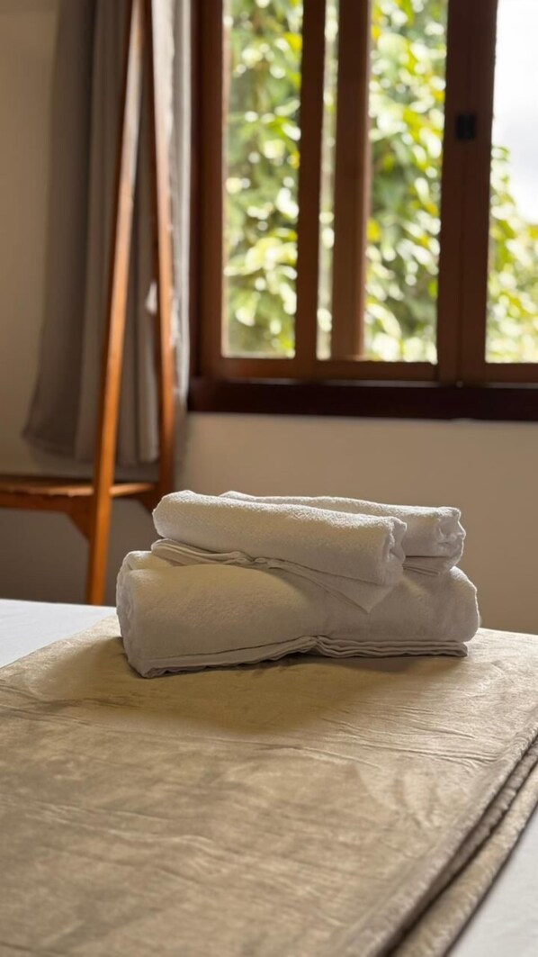 A neatly folded white towel on a bed with a wooden headboard, a window with vertical blinds, and a painting on the wall. - Go Hotelaria (Búzios)