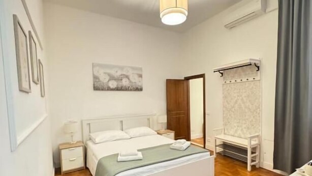 A bedroom with a bed, bedside tables, a desk, a chair, a hanging light fixture, and a painting on the wall.