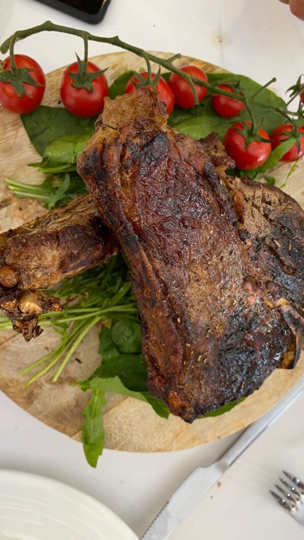 Pierna de cordero asada sobre tabla de madera con tomates cherry y verduras de hoja.