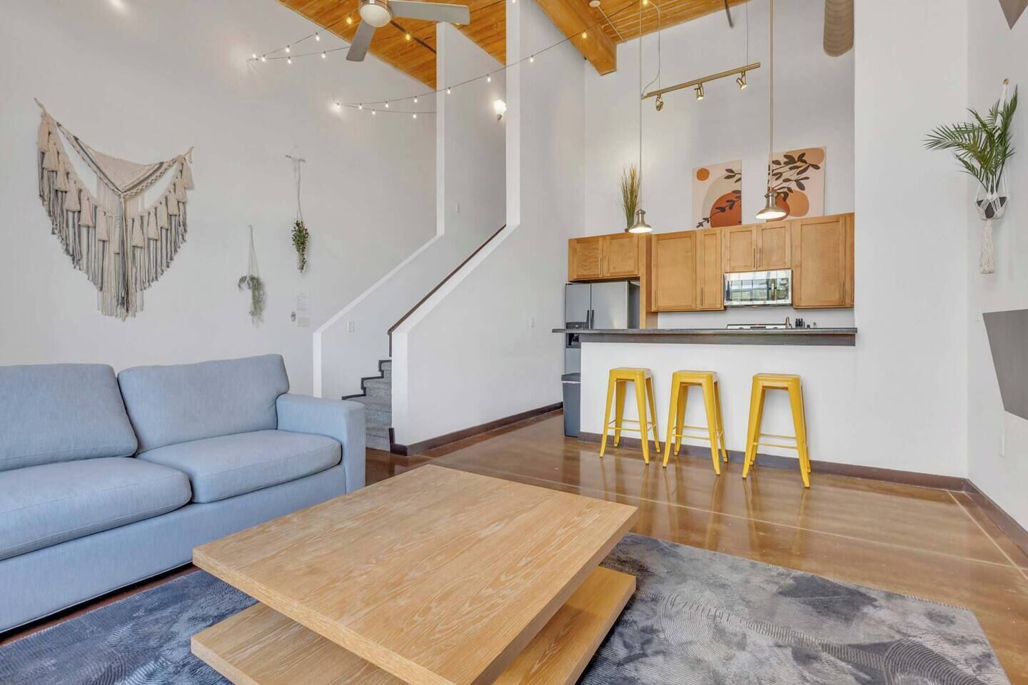 Un salon moderne avec un canapé, une table basse en bois et des tabourets jaunes. On aperçoit, au fond, une cuisine avec des meubles en bois et un escalier.