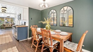 Een eethoek met een houten tafel, stoelen en een vaas met gedroogde planten. Achtergrond: een keuken met witte kasten en een fornuis.