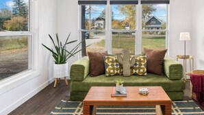 Een woonkamer met een groene bank, gestreepte kussens, een houten salontafel, een potplant en een uitzicht op huizen door grote ramen.