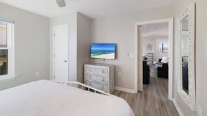 A bedroom with a bed, a dresser, a TV on the wall, and a hallway leading to a living area. - Beach Boyd's by Liquid Life (Gulf Shores)