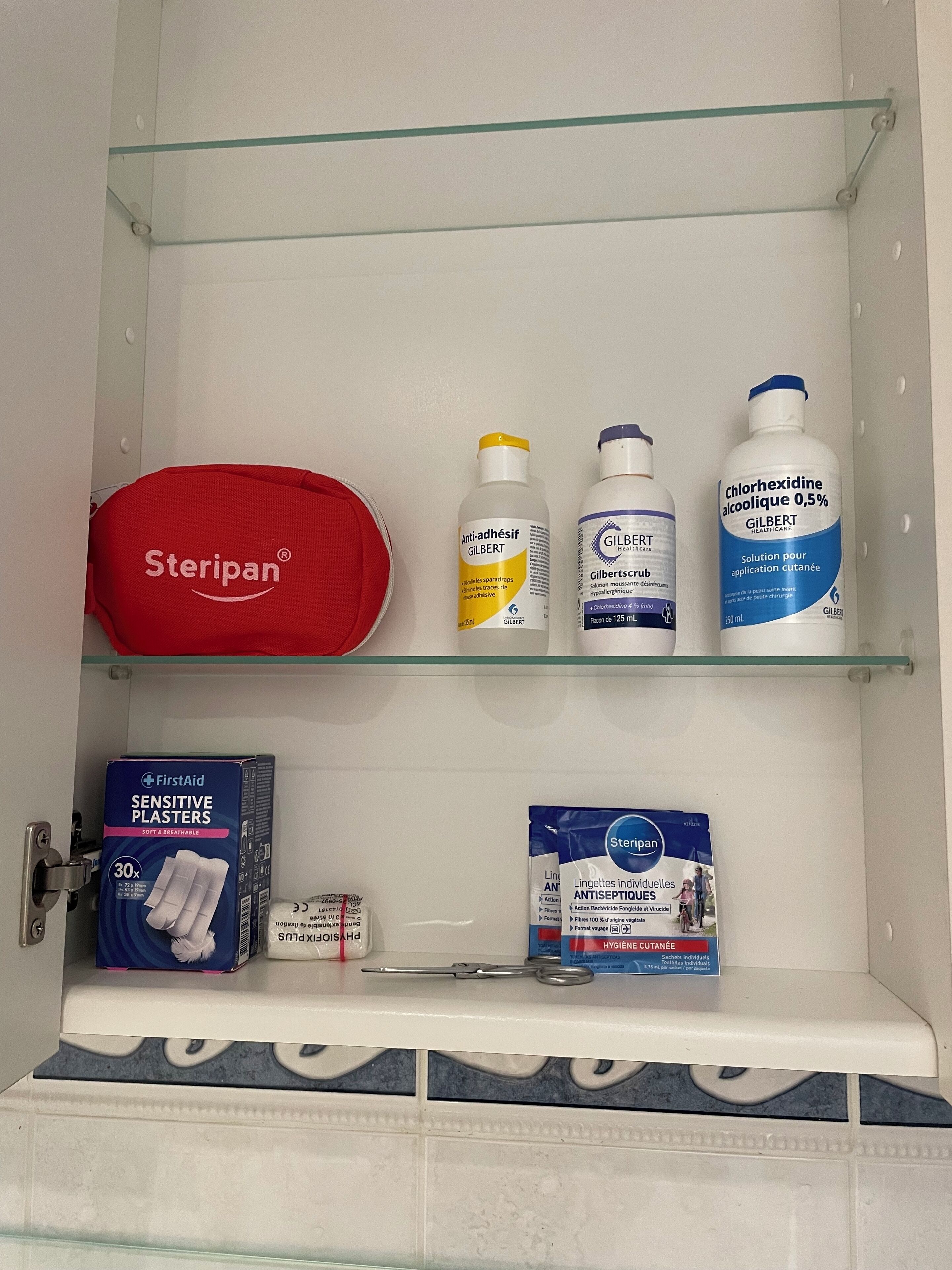 Un estante de baño con suministros médicos, incluyendo un Steripak rojo, dos botellas de solución antiséptica Gilbert, una caja de tiritas sensibles FirstAid y un paquete de toallitas individuales Steripak.