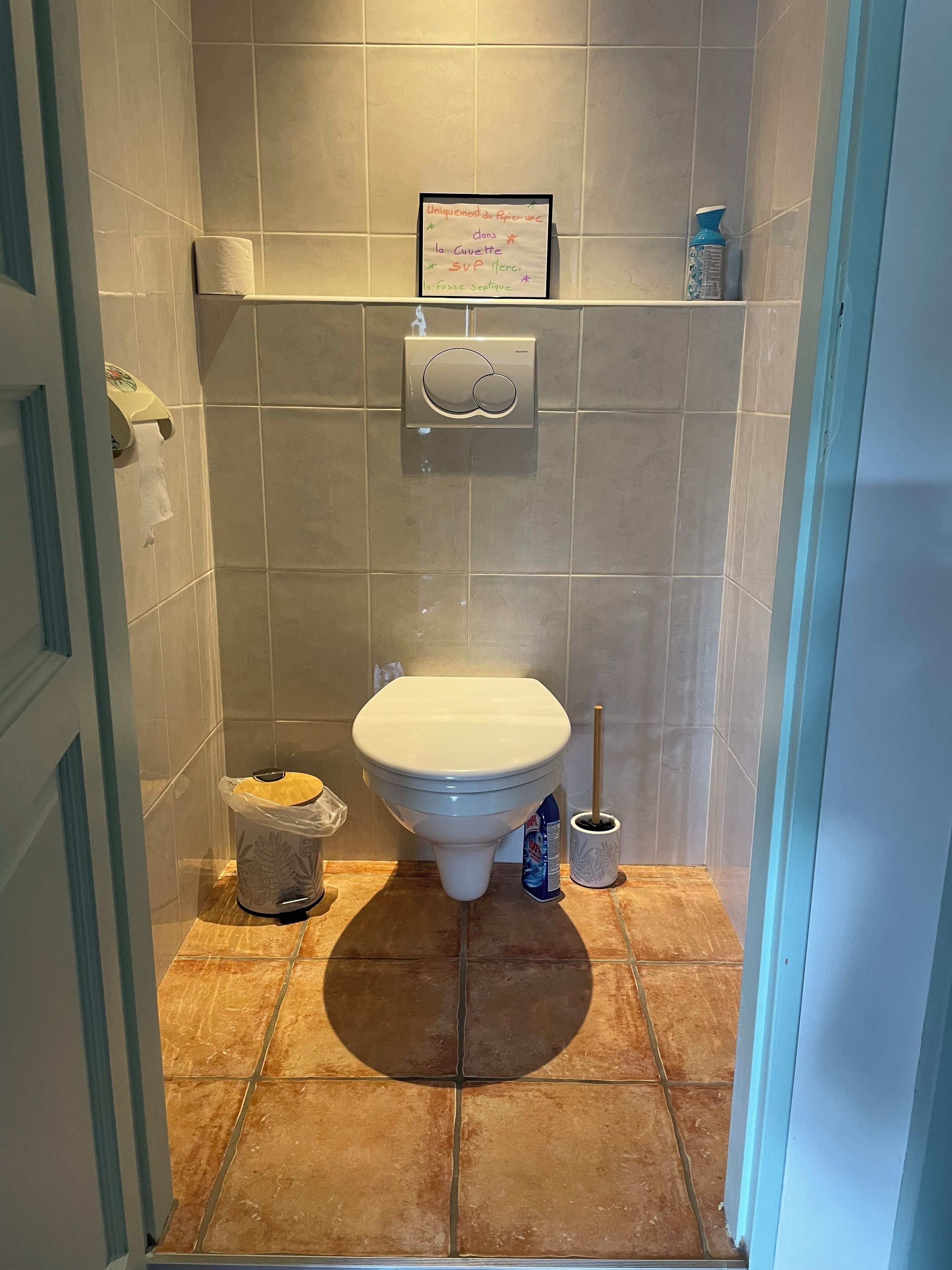 Ein Badezimmer mit Toilette, Papierhandtuchspender, Bidet und einer kleinen Ablage mit einem gerahmten Zettel.