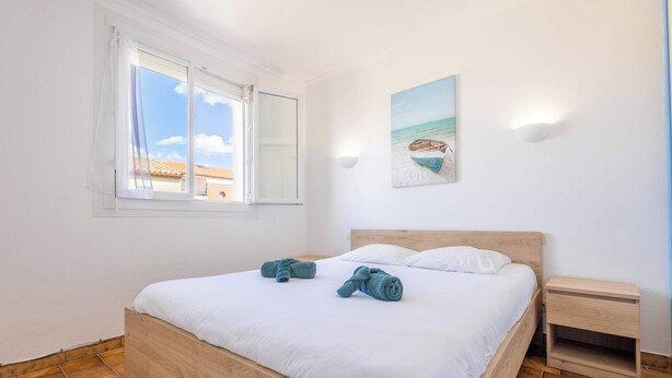 A bedroom with a wooden bed, white linens, a painting of a beach scene, a nightstand, and a window with curtains. - Le Repos Marin (Valras-Plage)