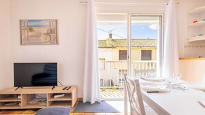 A room with a TV, a dining table set for two, a window with curtains, and a view of buildings outside. - Le Repos Marin (Valras-Plage)
