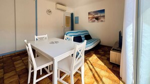 A room with a white table and chairs, a striped sofa, a painting on the wall, and a window with curtains. - Le Repos Marin (Valras-Plage)