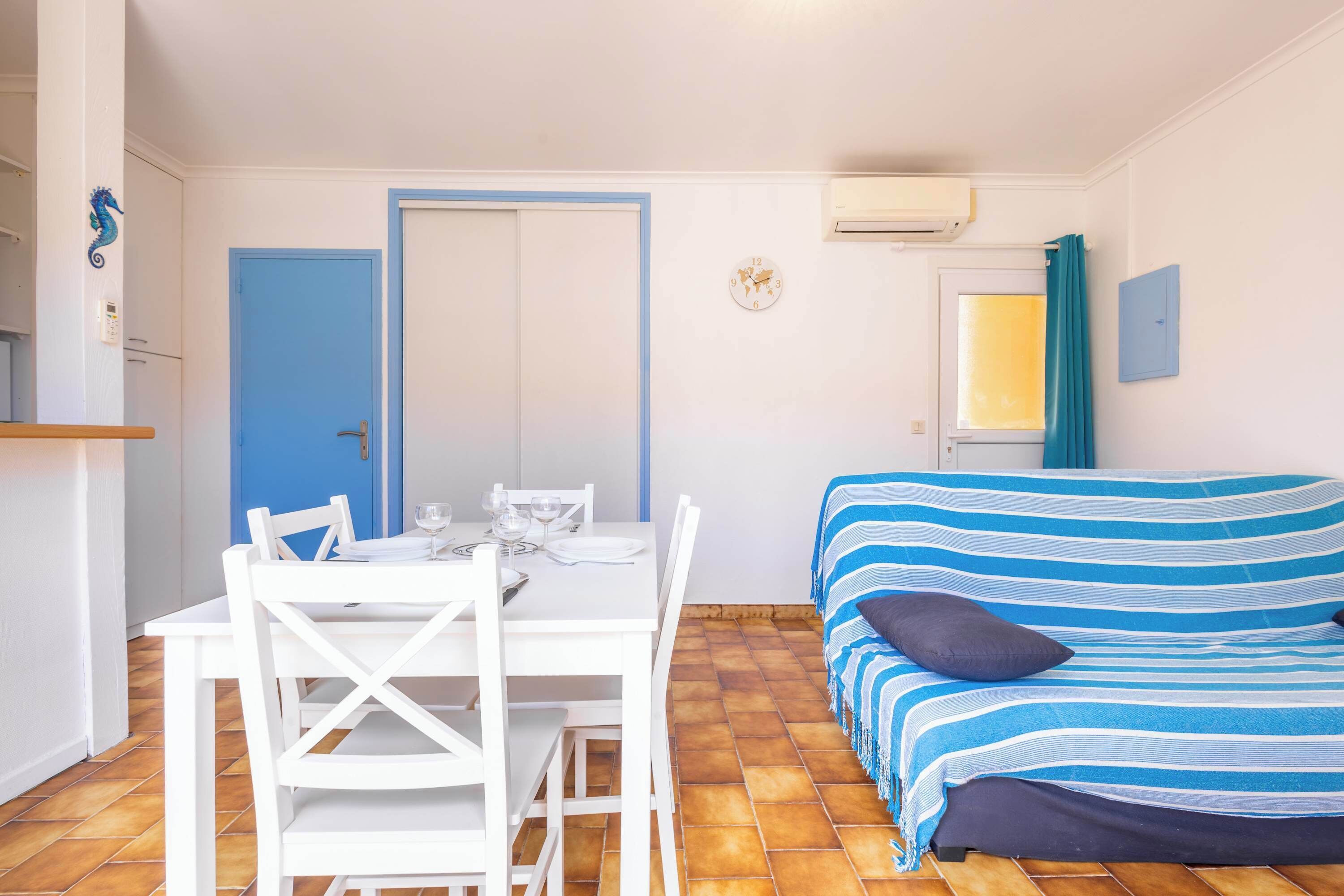 A room with a dining table set for two, a bed with striped bedding, a blue door, and a window with curtains.