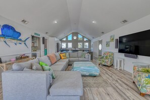 A spacious living room with a large sectional sofa, a flat-screen TV, and a dining area with a kitchen in the background. - Five Bedroom Home with a Private Pool on the Channel and Pet Friendly (Garden City Beach)