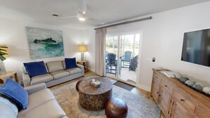 Sala de estar com sofá, mesa de centro e televisão. Há uma porta de vidro deslizante que dá para uma área externa com cadeiras.